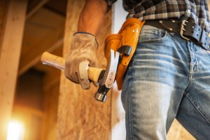 A skilled carpenter holds a hammer while wearing gloves and a tool belt, standing in a construction area illuminated by warm evening light.